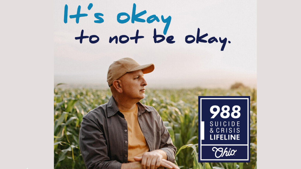 A middle-aged male in a khaki baseball cap looks across a field of corn. Blue text reads "It's okay to not be okay" the Ohio 988 Suicide & Crisis Lifeline logo is in the bottom right corner.